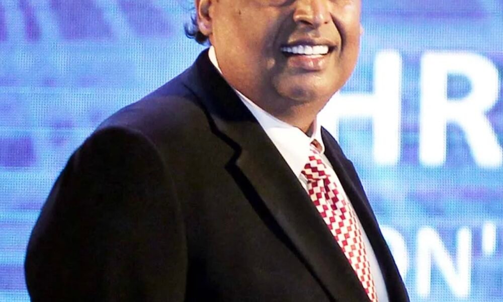Mukesh Ambani in a dark suit with a patterned tie standing in front of a blue background